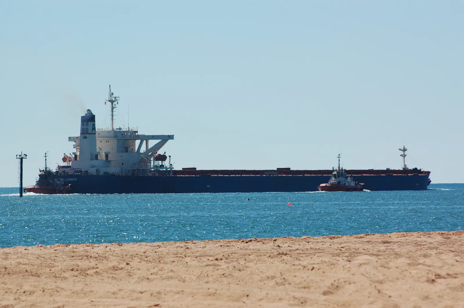 A loaded bulk ore carrier leaving Port Hedland with a tug alongside, seen from the beach